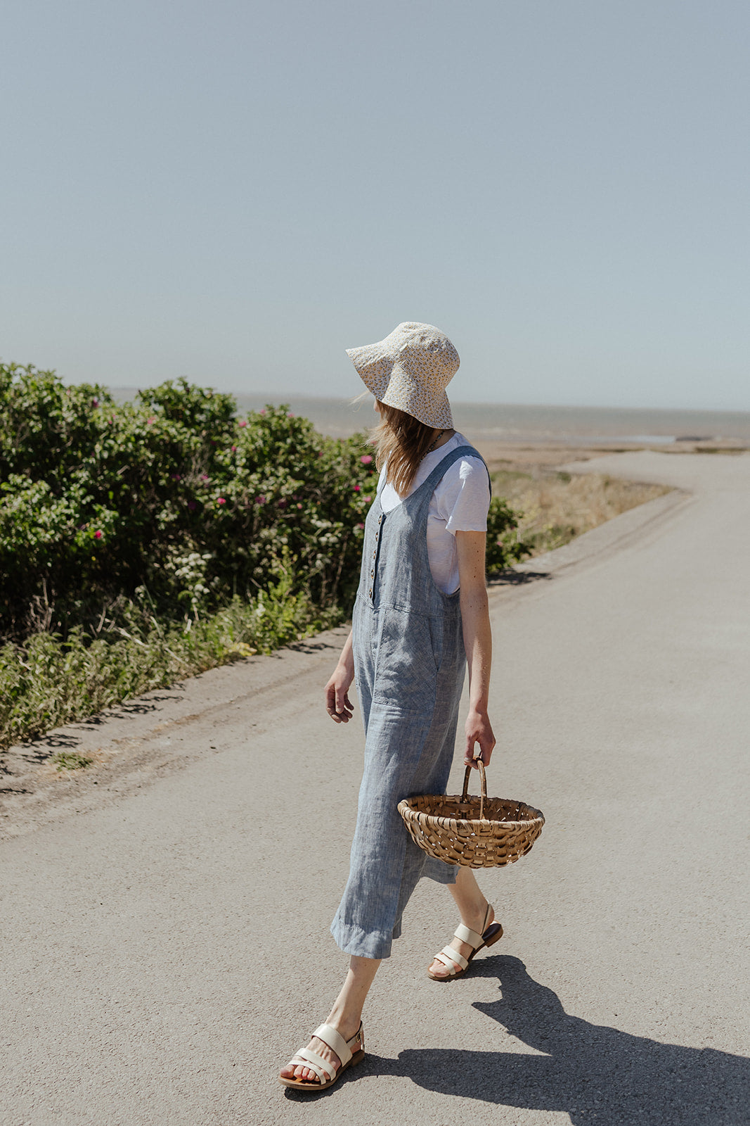 Woman in linen dungarees and white hat walking on a road with greenery and a clear sky