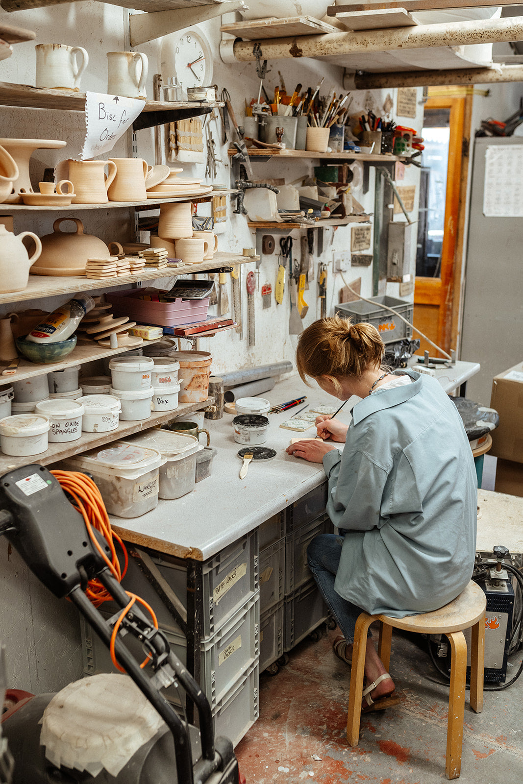 Person working in a pottery studio with shelves of clay items and tools.
